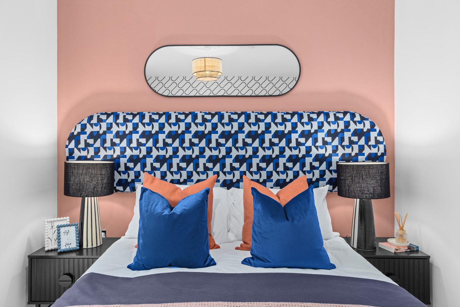 Close up of white bed with pink accent wall and blue/pink cushions.
