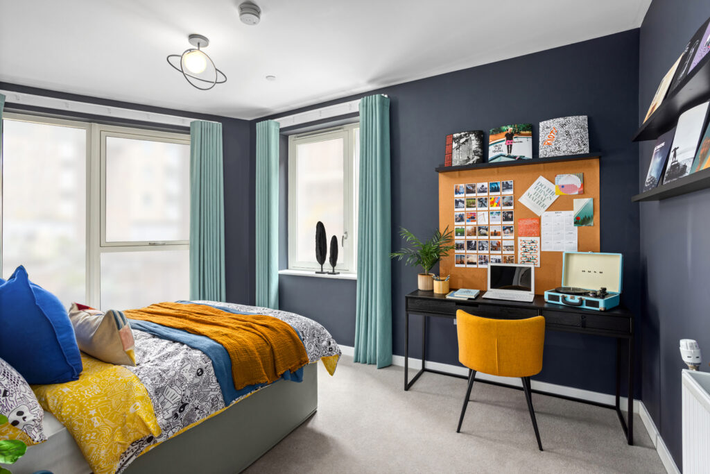 Bedroom with dark walls, bed with cushions and bedding and study desk with mustard yellow chair